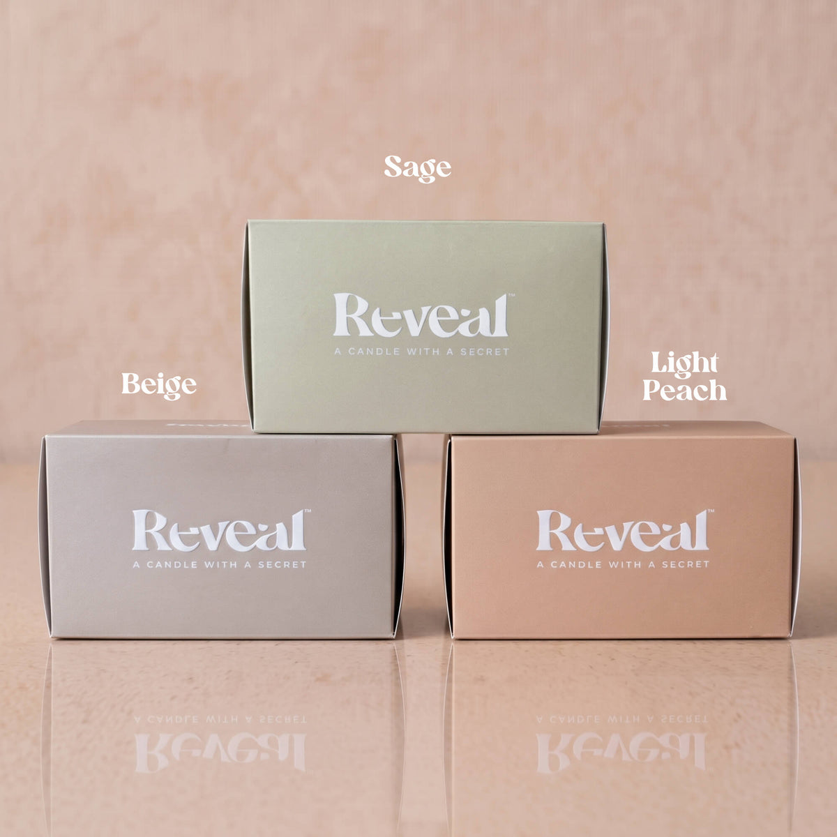 Three rectangular product boxes for Reveal candles are arranged in a pyramid on a reflective surface against a soft peach background. The top box is sage green and labeled 'Sage'; the bottom left is beige and labeled 'Beige'; the bottom right is light peach and labeled 'Light Peach'.