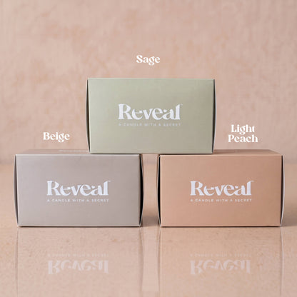 Three rectangular product boxes for Reveal candles are arranged in a pyramid on a reflective surface against a soft peach background. The top box is sage green and labeled 'Sage'; the bottom left is beige and labeled 'Beige'; the bottom right is light peach and labeled 'Light Peach'.